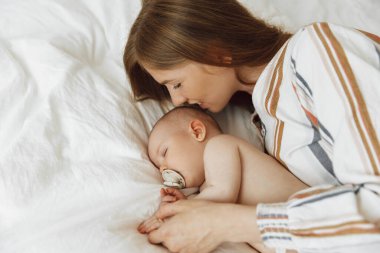 Close up portrait of a beautiful young mother kissing her newborn baby sleeping on the bed at home. Smiling mother and baby are lying on the bed at home. The baby sleeps comfortably with the safety and protection of the mother. Health care, love, lif