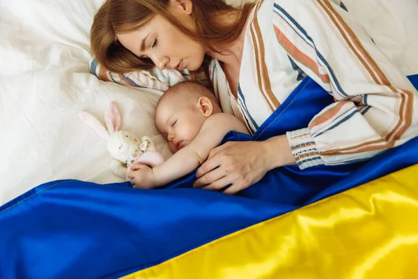 Close-up portrait of a mother kissing her newborn baby sleeping on the bed at home covered with the Blue and yellow flag of Ukraine. The baby sleeps with the safety and protection of the mother. Family, unity, support. Ukrainians against war, Mother\'