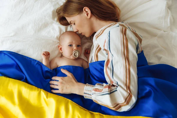 Close-up portrait of a mother kissing her newborn baby sleeping on the bed at home covered with the Blue and yellow flag of Ukraine. The baby sleeps with the safety and protection of the mother. Family, unity, support. Ukrainians against war, Mother\'