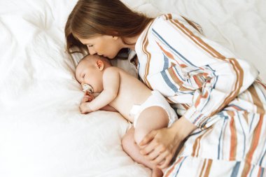 Close up portrait of a beautiful young mother kissing her newborn baby sleeping on the bed at home. Smiling mother and baby are lying on the bed at home. The baby sleeps comfortably with the safety and protection of the mother. Health care, love, lif
