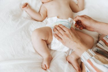 Side view portrait of a mother changing her baby's diaper on the bed, one year old baby concept. The mother changed the baby's diaper in bed, the woman puts on the baby's diaper. Close up, with copy space