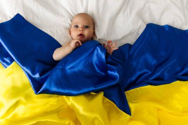 Portrait of a baby wrapped in the national blue and yellow flag of Ukraine, lying on the bed. Baby with Ukraine flag heart. photo with place for text. Children of war
