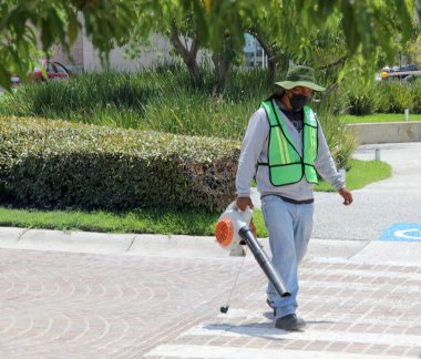 Maskeli bir adam, yüksek görüş mesafesi olan bir güvenlik yeleği ve şapkası var. Güneşte bir yaprak üfleyicisi ile sokakları temizliyor.