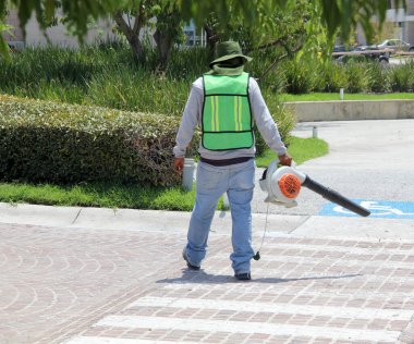 Maskeli bir adam, yüksek görüş mesafesi olan bir güvenlik yeleği ve şapkası var. Güneşte bir yaprak üfleyicisi ile sokakları temizliyor.