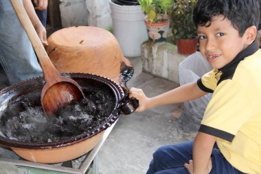 Zavallı Latin çocuk, düşük gelirli evinin arka bahçesinde kil çömlek ve tahta kaşıkla yemek pişirmeye yardım ediyor.