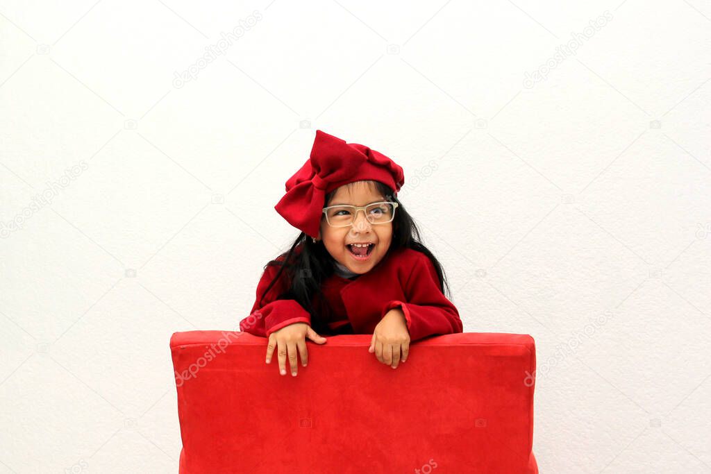 Little Latin girl with glasses dressed very elegantly for a party with a red hat is very happy and ready to celebrate in a red chair where the celebration will take place such as Christmas, birthday, new year, thanksgiving, love day