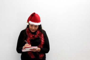 Şapkası ve Noel çelengi olan Latin yetişkin kadın, tebrik, aşk ya da dilek listesi mesajı vermek için pankart ve boş not defteri gösteriyor.