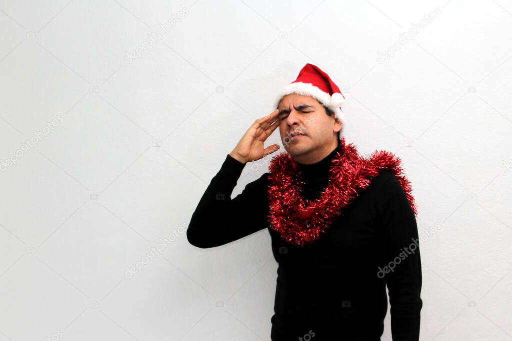 Dark-haired Latin adult man with hat and Christmas garland shows his anger, disgust and sadness for the arrival of December, he does not like Christmas