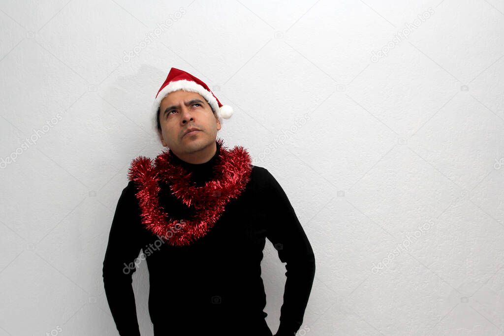 Dark-haired Latin adult man with hat and Christmas garland shows his anger, disgust and sadness for the arrival of December, he does not like Christmas