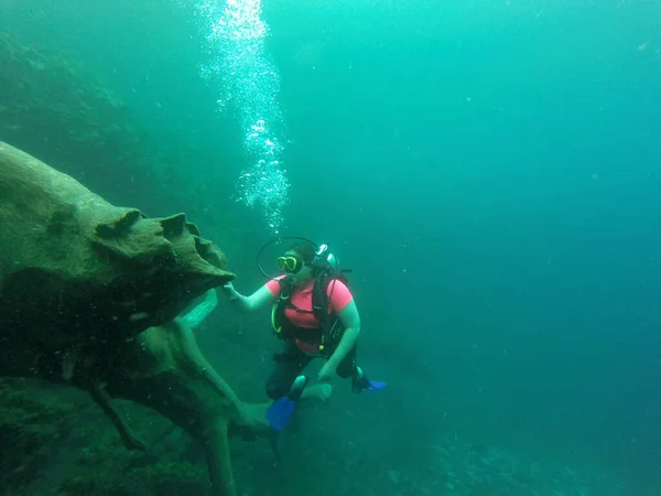 Young woman practices the sport scuba diving with oxygen tank equipment, visor, fins, relaxes and enjoys the bottom of the crystal clear water next to large branches and trunks