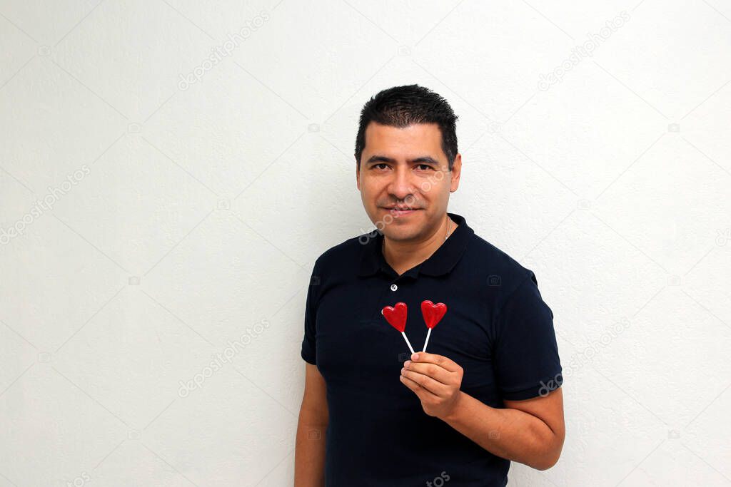 Latino adult man with heart-shaped candy lollipops shows his enthusiasm for the arrival of February and celebrate Valentine's Day of Love and Friendship with his partner, friends and family