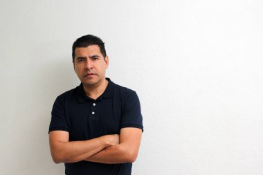 Kısa siyah saçlı, asul lacivert tişörtlü, beyaz bir duvarın önünde modellik yapan yetişkin bir Latin.