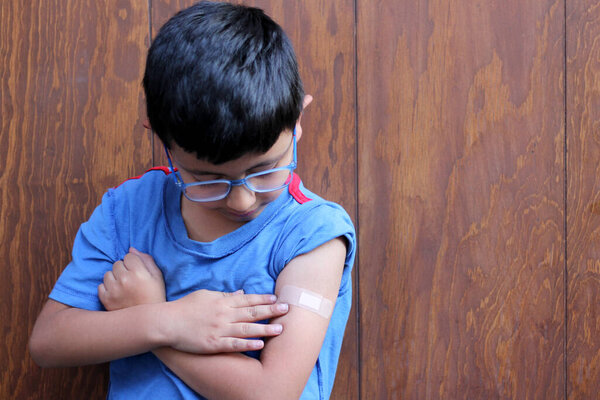 Маленький 6-летний латиноамериканец в очках и голубой рубашке показывает руку с бинтом, потому что он только что был вакцинирован против Ковида-19 в новой нормальности из-за пандемии коронавируса
