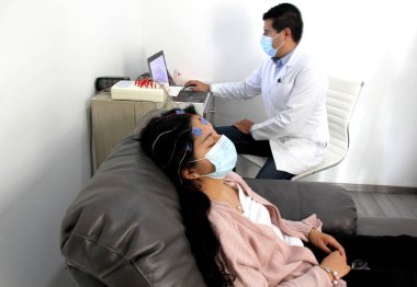 Tıbbi muayenelerde koruyucu maskeli Latin kadın ve doktor covid-19 vakalarında elektroensefalogram ve beyin haritalama yapıyorlar.