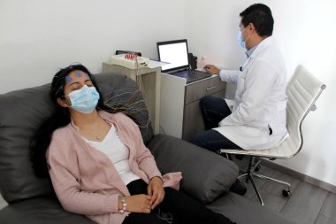 Tıbbi muayenelerde koruyucu maskeli Latin kadın ve doktor covid-19 vakalarında elektroensefalogram ve beyin haritalama yapıyorlar.