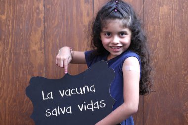 Kıvırcık saçlı Latin küçük kız, kolunun yakın zamanda Covid-19 'a karşı aşılandığını gösteriyor. Üzerinde 