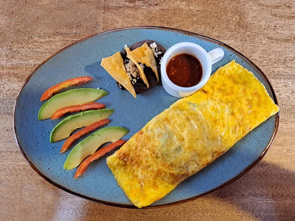 Meksika usulü omlet kahvaltısı, mantarla doldurulmuş avokado, domates, kırmızı sos, peynirli fasulye ve ahşap bir masanın yanındaki pencerenin yanında tortilla cipsi.