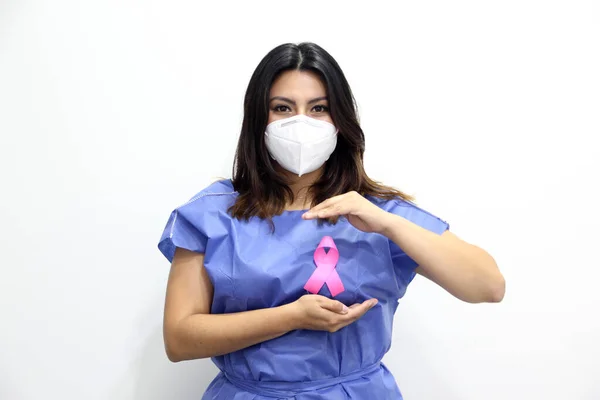 Latin woman with protection mask, disposable blue surgical gown and pink ribbon for campaign against breast cancer