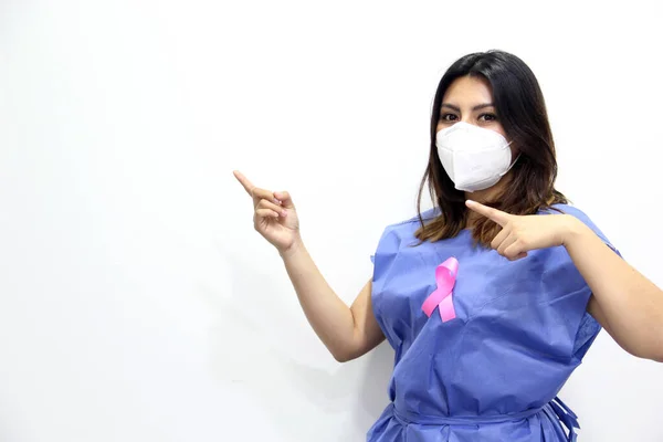 Latin woman with protection mask, disposable blue surgical gown and pink ribbon for campaign against breast cancer