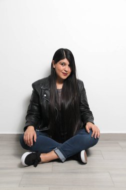 Mutlu, şişman ve gururlu Latin yetişkin kadın uzun düz, siyah ve parlak saçlarını beyaz arka planda deri ceket ve siyah tenis ayakkabılarıyla gösteriyor.