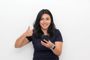 Zafer pozisyonunda cep telefonuyla izole edilmiş beyaz arka planı olan genç Latin kadın mutlu ve heyecanlı görünüyor.