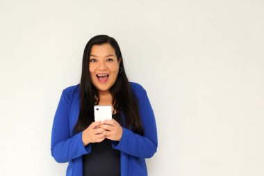 Düz siyah saçlı ve mavi ceketli Latin yetişkin bir kadın cep telefonundan çok heyecanlı ve mutlu bir şekilde internetten alışveriş yapıyor.