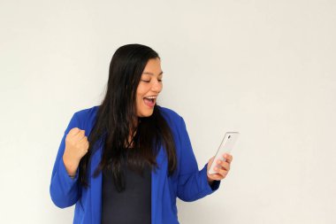 Düz siyah saçlı ve mavi ceketli Latin yetişkin bir kadın cep telefonundan çok heyecanlı ve mutlu bir şekilde internetten alışveriş yapıyor.