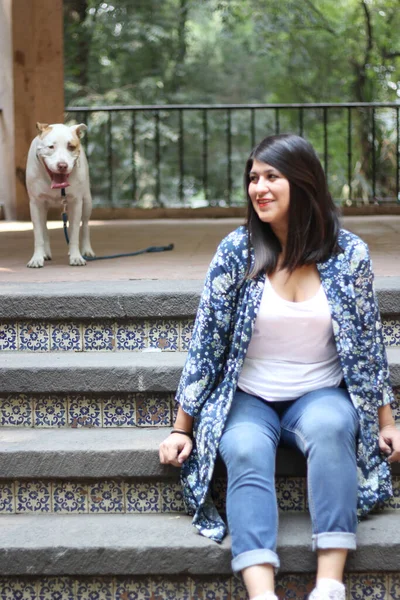 Latin adult woman relaxes sitting in a park kiosk accompanied by her white pitbull dog does not grab the leash and picks up waste