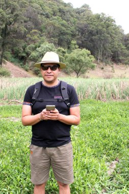 Şapkalı, şortlu ve güneş gözlüklü Latin yetişkin bir adam ormanın ortasında cep telefonuyla mesaj gönderiyor ve alıyor. 