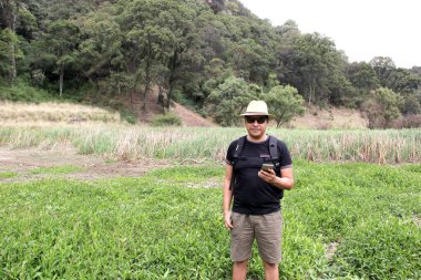 Şapkalı, şortlu ve güneş gözlüklü Latin yetişkin bir adam ormanın ortasında cep telefonuyla mesaj gönderiyor ve alıyor. 