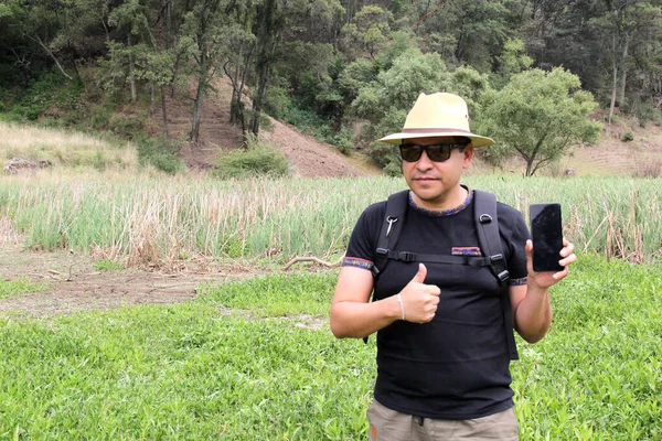 Şapkalı, şortlu ve güneş gözlüklü Latin yetişkin bir adam ormanın ortasında cep telefonuyla mesaj gönderiyor ve alıyor. 