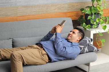 Latin yetişkin bir adam cep telefonunu bir koltukta kullanır. Arama yapar, video görüşmeleri yapar, mesajlar gönderir, internetten satın alır, sosyal ağlar kullanır ve aşkı arar.