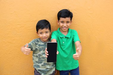 Yoksulluk içinde yaşayan iki siyah saçlı Latin erkek çocuk oyun oynamak için cep telefonlarını kullanıyor ve oyun için zafer işareti yapmak için heyecanlılar.