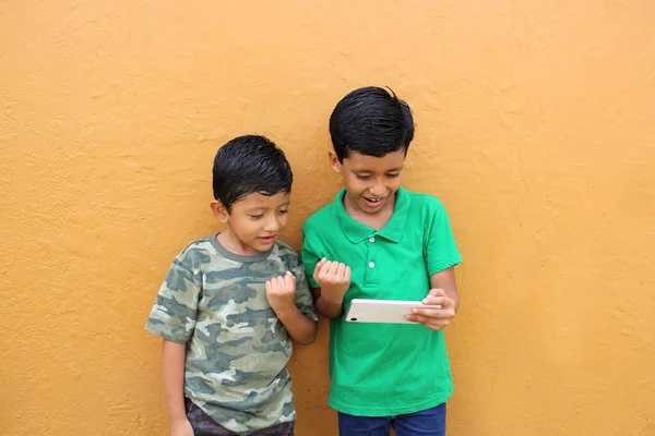 Two dark-haired Latino male children living in poverty use their cell phones to play games and are excited to make a victory sign for the game