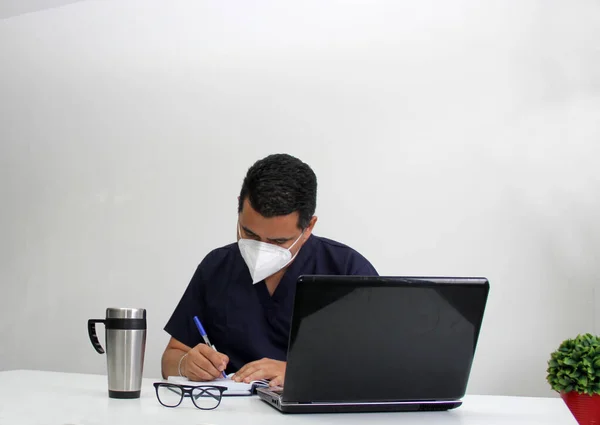 Latino medical specialist doctor works in his office, writes prescriptions with clinical face mask next to his laptop, glasses and coffee stressed and exhausted