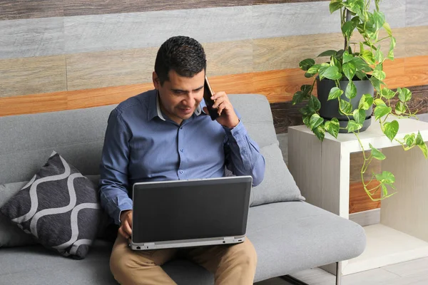 Single Latino adult man does home office in his living room with laptop and cell phone, creates reports, video calls, overtime in the new normal without an office