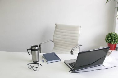 Rahat koltuğu, bitkileri olan beyaz boş bir ofis, işe başlamaya hazır dizüstü bilgisayar, cep telefonu, defter, bardaklar ve termosta kahve