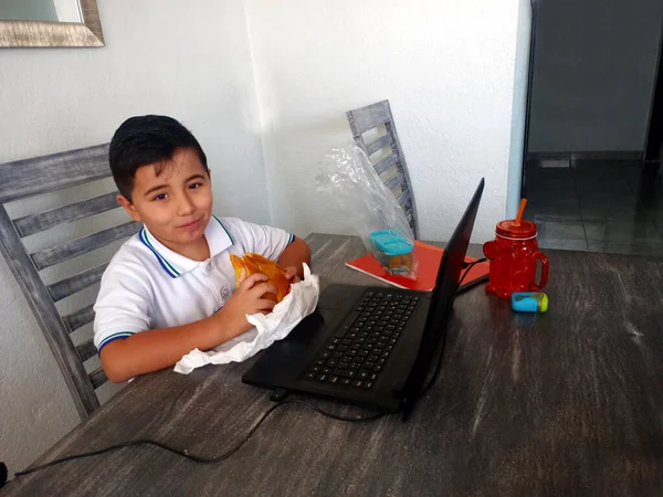 Boy does home school with his laptop and uniform in the dining room and eats a sandwich at break time and rest inside his house in the new normal