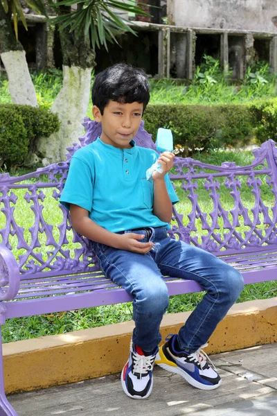 Poor Latino dark-haired boy with a blue t-shirt sitting on a park bench eating an ice pop because of the heat wave and cooling off in poverty by affecting his teeth with sweets and sugar