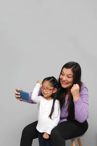 Single divorced Latina mom and daughter use cell phone to video call, take selfies, play video games, and shop online