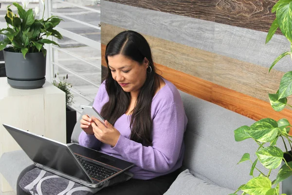 Latin adult woman sitting on a sofa works with her laptop and cell phone outside the office doing home office talks on video call and sends messages under pressure and stress