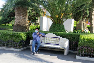 Koruyucu maskeli latin adam park bankında oturuyor. Yeni normal Coffid-19.