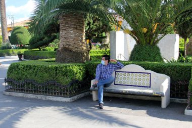 Koruyucu maskeli latin adam park bankında oturuyor. Yeni normal Coffid-19.