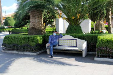 Koruyucu maskeli latin adam park bankında oturuyor. Yeni normal Coffid-19.