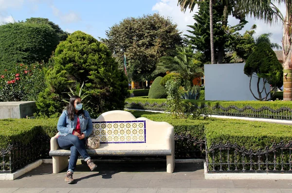 Siyah saçlı, koruyucu maskeli, parktaki bankta oturan Latin bir kadın.