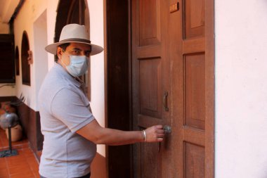 Koruyucu maskeli Latin adam klinik ve şapka takıyor, içeride, odasının ahşap kapısını açıyor.