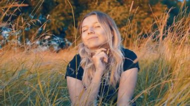 Sonbaharda sarı çimenlerde oturan uzun sarı saçlı güzel bir kızın duygusal portresi. Sonbaharın başlarında günbatımında doğanın arka planına karşı. Duygular ve seçici bir gülümseme..