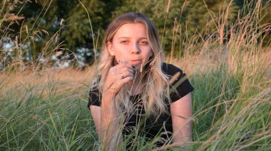 Güzel, uzun saçlı, gülümseyen sarışın bir kızın portresi. Sonbahar doğasının arka planına karşı çimlerde oturuyor. Gün batımındaki duygu kavramı..