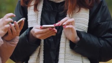 Sarı bir sonbahar parkında iki genç kız yakın planda telefonlarına mesaj yazıyorlar..