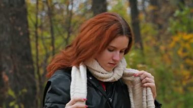 Beyaz eşarplı genç bir kız, kırmızı altın sarısı saçlarını büyük bir planla sonbahar sarı parkının arka planına göre ayarlıyor..